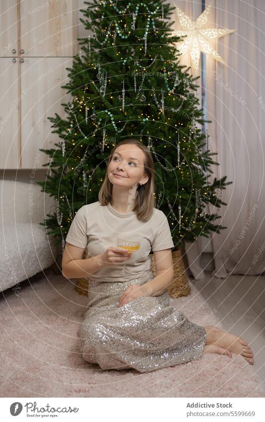 Joyful young girl sitting by a Christmas tree with star decoration and drinking a cocktail christmas festive season cheerful smile floor adorn celebrate holiday