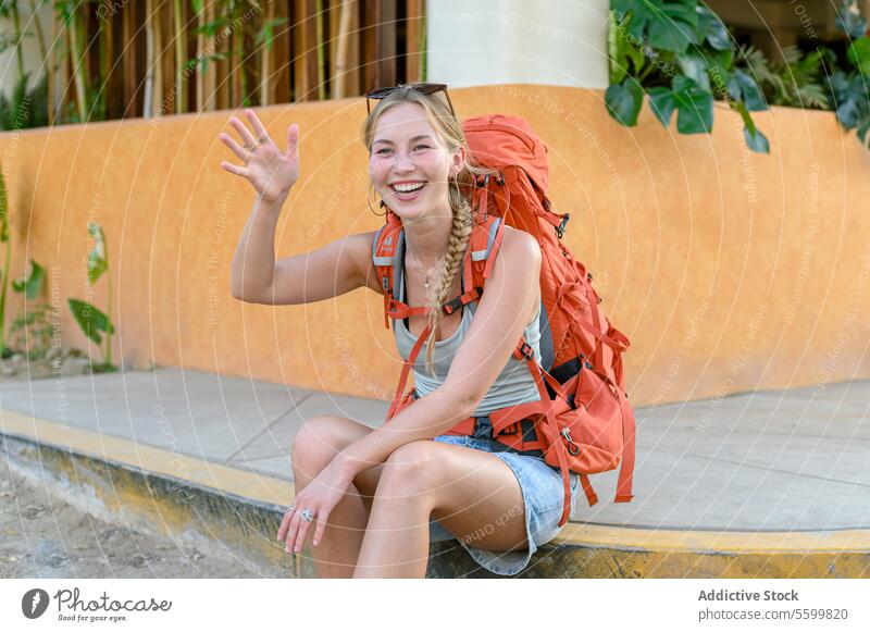 Cheerful young adult woman tourist with blond long hair orange backpack casual clothes sitting on stone street border waving hand while waiting for friend and laughing against tropical background