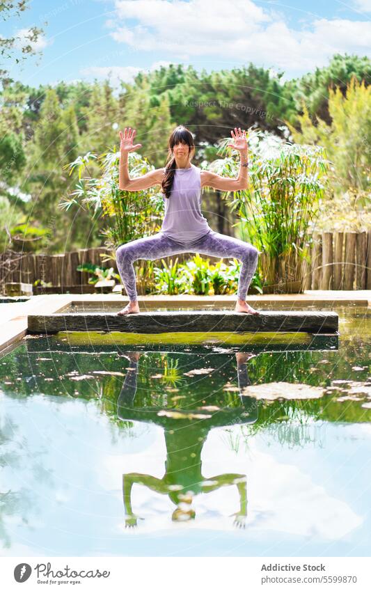 A woman practicing the yoga pose Goddess with Cactus Arms on a tree trunk over a pool, Vertical shot aesthetic arms balance bridge cactus calmness concentration