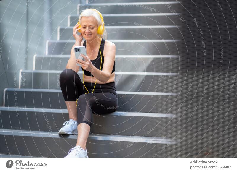 Senior Woman Enjoying Music on Smartphone Outdoors in workout attire woman senior smartphone headphones music outdoor stairs sitting active lifestyle healthy