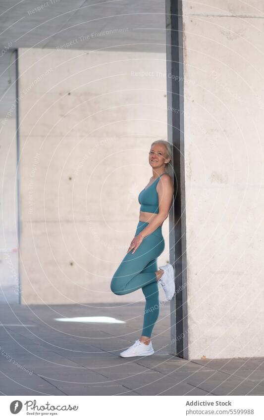 Fit senior woman in activewear leaning against a wall fitness smiling teal break urban concrete exercise wellness health mature sporty lifestyle relaxed happy