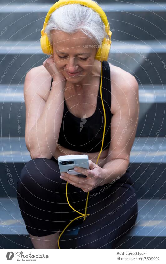 Senior woman enjoying music on headphones outdoors in workout attire. senior smartphone steps silver hair smiling leisure lifestyle relaxation technology