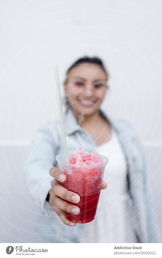 A young Hispanic woman is smiling while drinking a Mexican cherry slushy outdoors sunglasses black hair blonde highlights earring denim jacket red straw ice