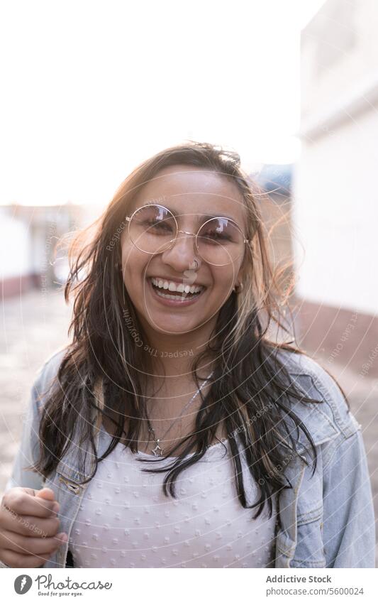 A young Hispanic woman with sunglasses is smiling posing in the street during the sunset black hair blonde highlights necklace piercing denim jacket smile laugh