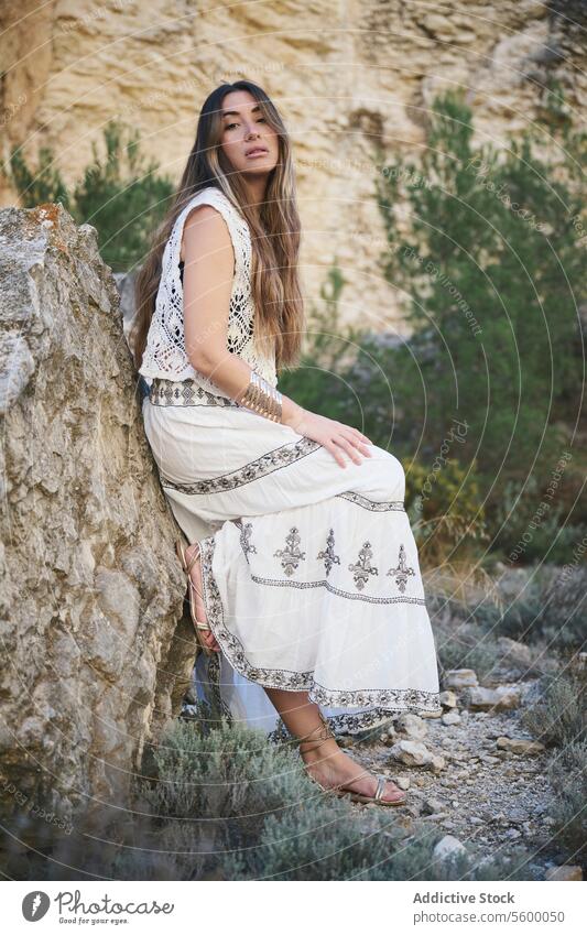 Woman in an embroidered dress next to a rock woman white bohemian landscape natural outdoor serene hair flow lean summer style fashion elegance calm pattern