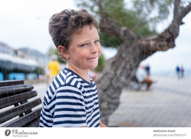 A child in a striped shirt gives a playful glance over his shoulder near a tree-lined promenade sea outdoor day casual leisure coastline vacation young boy