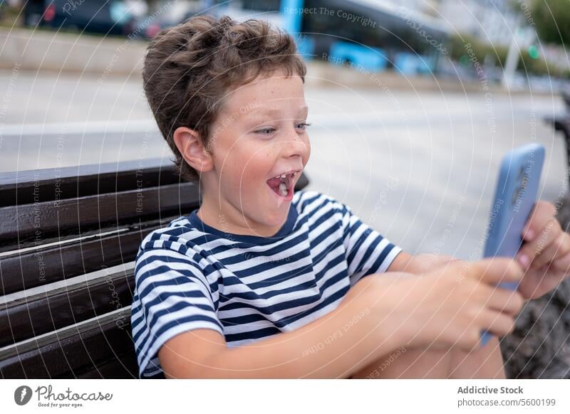Side view of young amazed boy sitting on a bench at the seaside happily using a smartphone happy technology outdoor child leisure coast port promenade casual