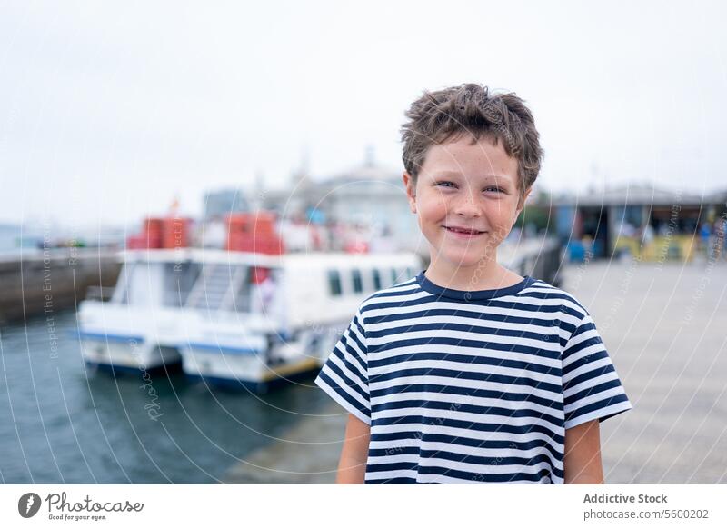 Cheerful young child in a striped t-shirt smiles at the camera with the sea and boats in the background during summer vacation happy striped shirt joy cheerful