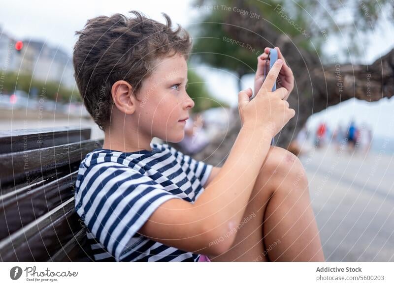 Side view of young boy sitting on a bench at the seaside happily using a smartphone happy technology outdoor child leisure coast port promenade casual summer