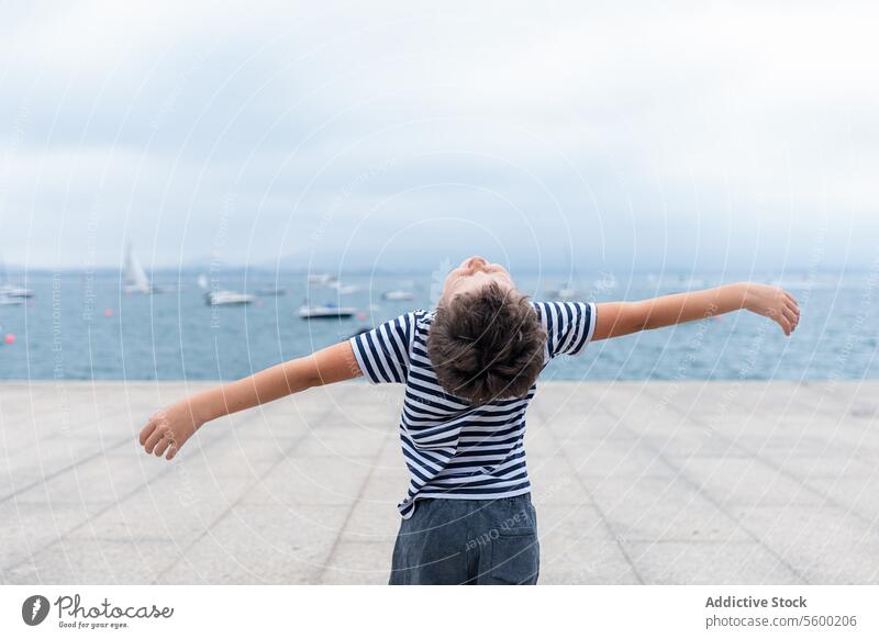 Child with outstretched arms stands by the sea, expressing freedom and joy.Title: Joyful child by the sea arms outstretched striped shirt horizon boats water