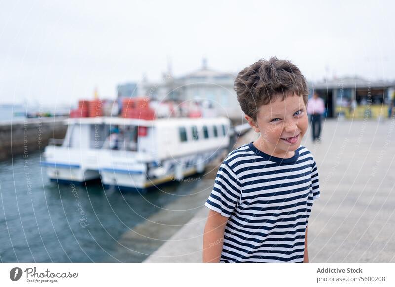 Cheerful young child in a striped t-shirt smiles at the camera while sticking tongue out with the sea and boats in the background during summer vacation happy