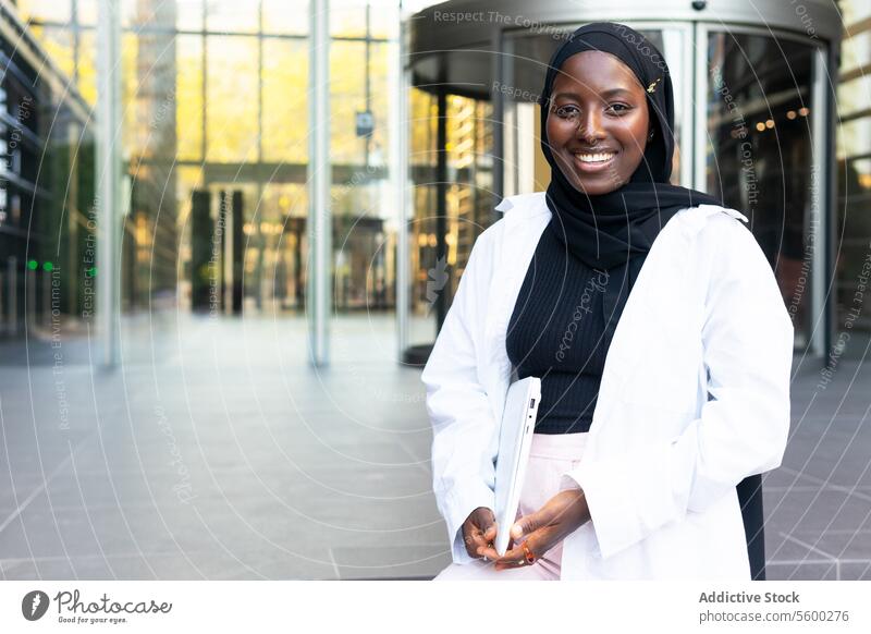 Cheerful Muslim African American female entrepreneur dressed in hijab and formals holding laptop and looking at camera while standing on street businesswoman