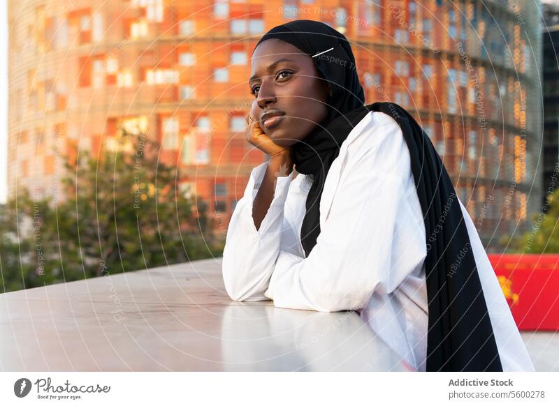 Side view of thoughtful African American Muslim businesswoman in black hijab touching chin and looking away while leaning on railing in city african american
