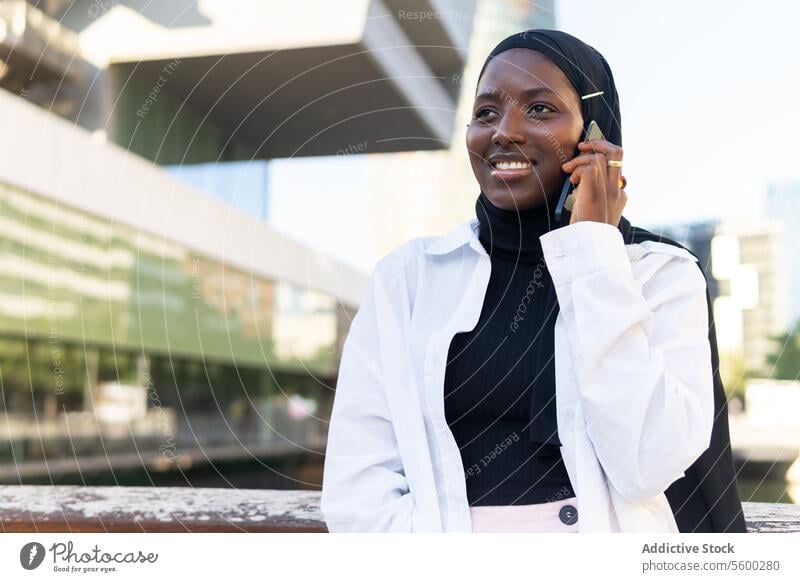 Happy woman in hijab talking over mobile phone in city businesswoman smartphone eyes closed cheerful modern building standing african american cellphone formal