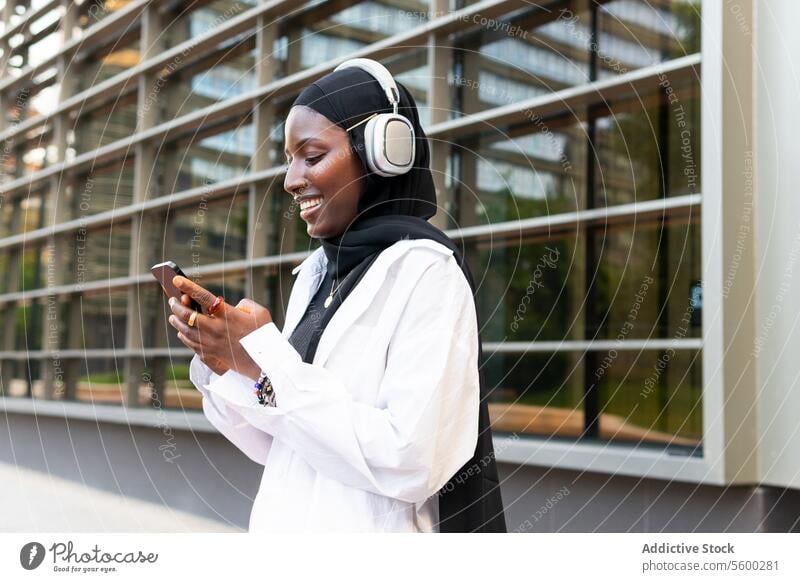 Happy woman with headphones and cellphone in city businesswoman headscarf listen music smartphone cheerful hijab smile muslim happy song mobile modern building