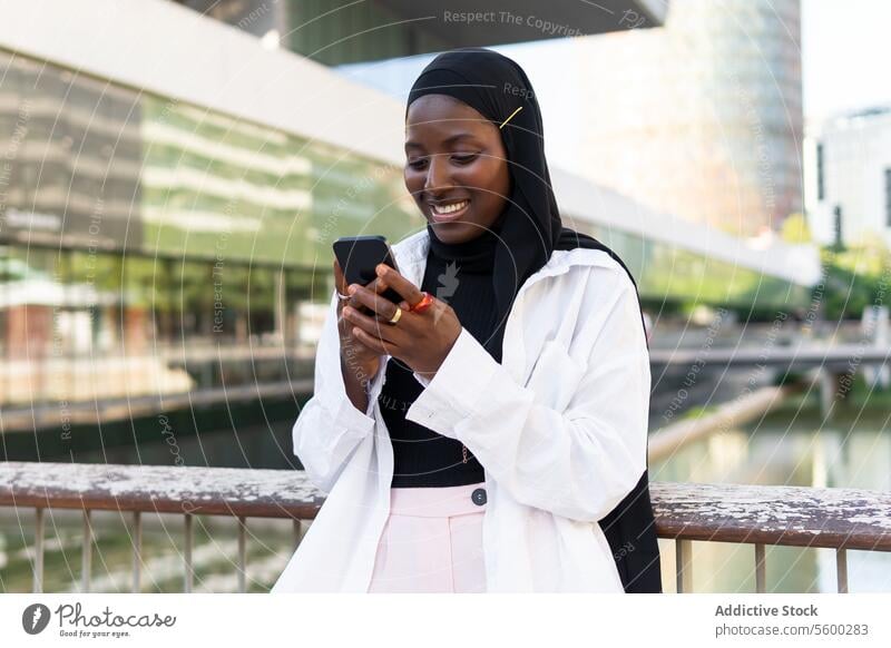 Cheerful African American young businesswoman in hijab browsing on smart phone while standing against modern building smartphone mobile phone african american