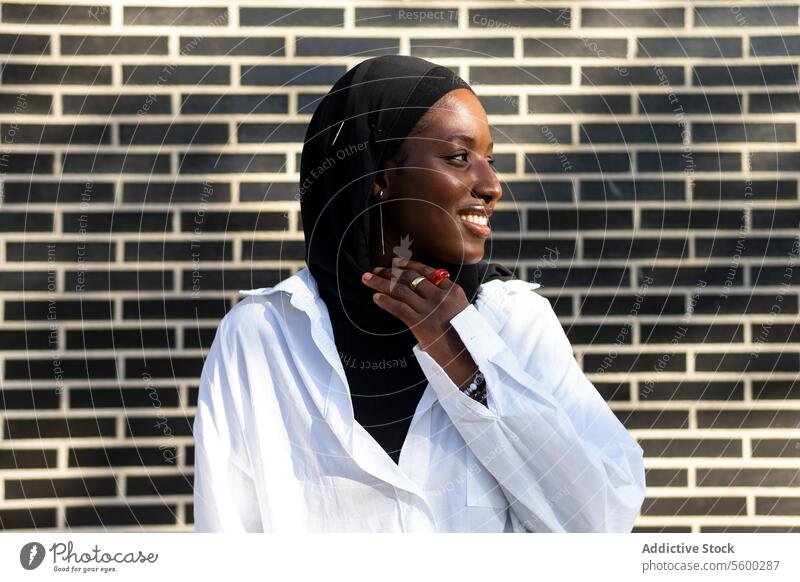 Portrait of confident smiling African American Muslim female entrepreneur in black hijab and white formal standing against brick wall in city businesswoman