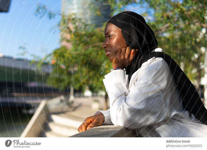 Side view of thoughtful African American Muslim businesswoman in black hijab touching chin and looking away while leaning on railing in city african american