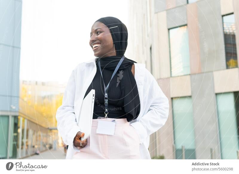 Cheerful Muslim African American female entrepreneur dressed in hijab and formals holding laptop and looking away while standing on street businesswoman muslim