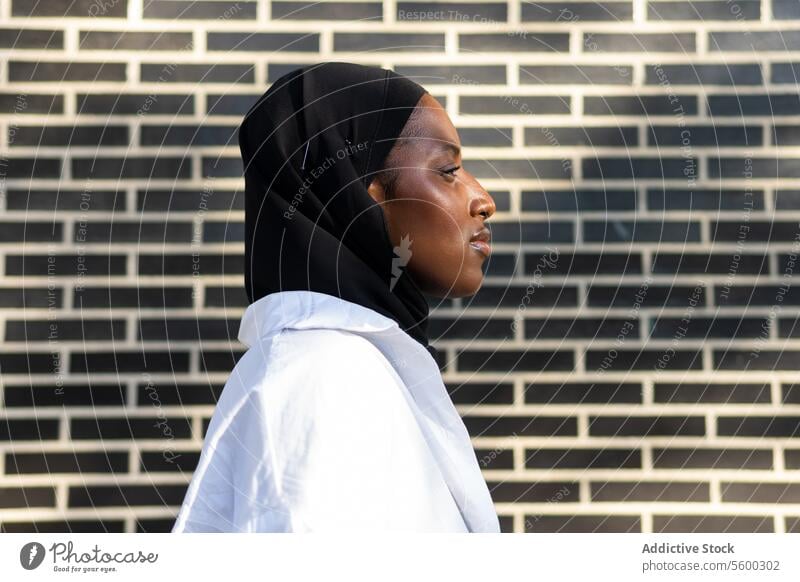 Portrait side view of confident African American Muslim female entrepreneur in black hijab and white formal standing against brick wall in city businesswoman