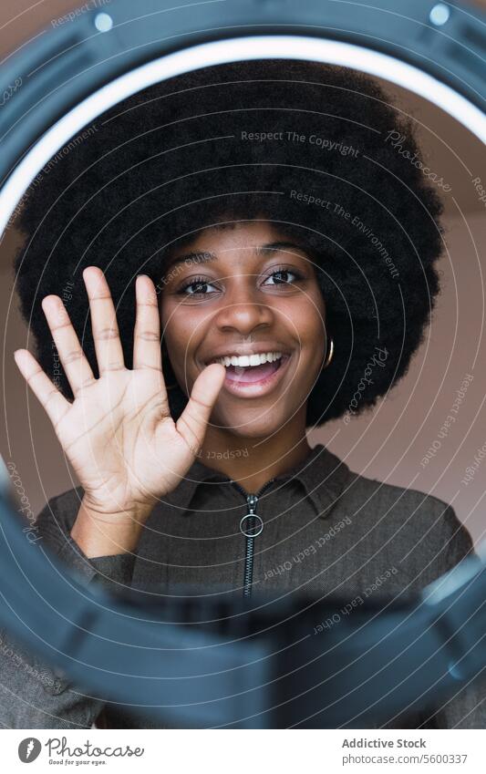 Content black woman waving hand though ring lamp vlog blogger wave hand greeting social media afro lighting smile hairstyle feminine appearance happy charming