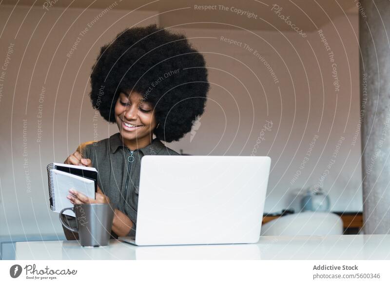 Cheerful black woman writing in notebook near laptop work freelance workplace take note online agenda schedule modern netbook home female write notepad