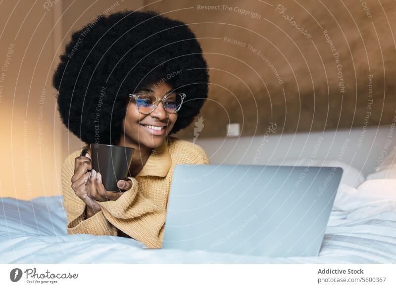 Smiling black woman with laptop and coffee in bed using rest relax weekend free time home female african american afro watch netbook movie comfort bedroom happy