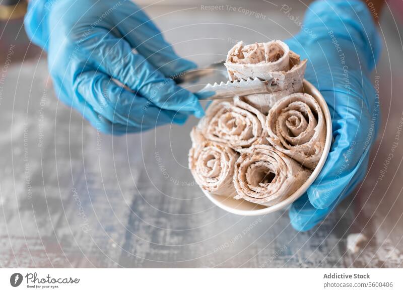 A worker is putting chocolate ice cream rolls in a tub using tongs and latex gloves hand rolled flat nut counter almond tray milk senior topping spatula mix