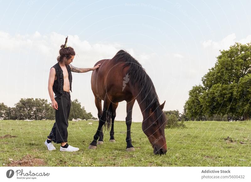 Man with tattoos and bare chest touching a horse in a green field in daylight man brown horse grazing pasture sunlit tree background nature outdoor explorer