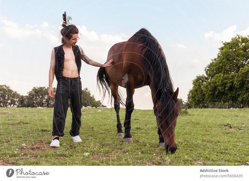 Man with tattoos and bare chest touching a horse in a green field in daylight man brown horse grazing pasture sunlit tree background nature outdoor explorer