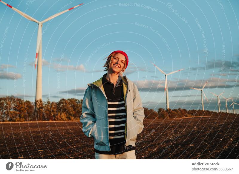 Smiling alternative woman looking at camera in denim jacket by wind turbines yellow beanie sustainable green energy cheerful young adult portrait dreadlocks