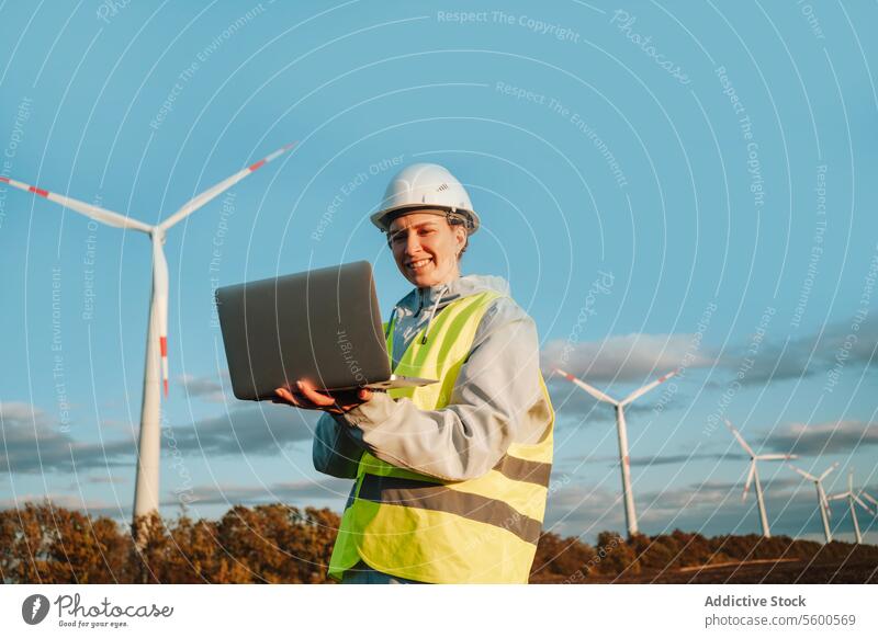 Engineer woman with laptop at wind farm during sunset engineer wind turbine safety gear renewable energy technology safety helmet smiling outdoors industry