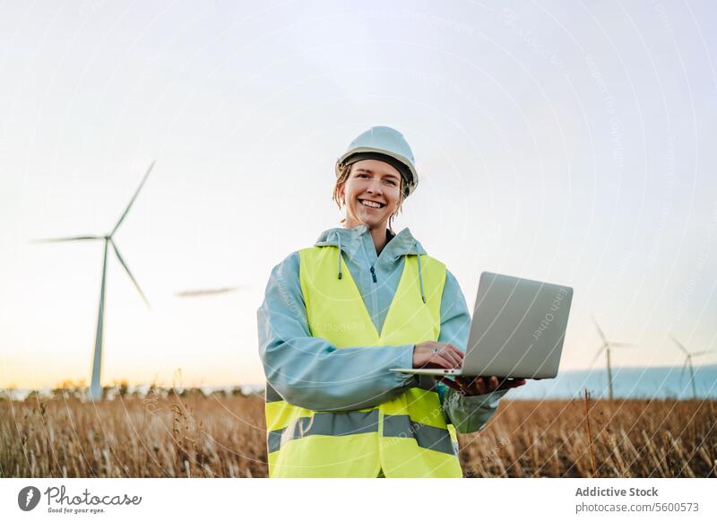 Engineer with laptop at wind farm during sunset engineer woman wind turbine sustainable energy technology field safety helmet high-visibility jacket environment