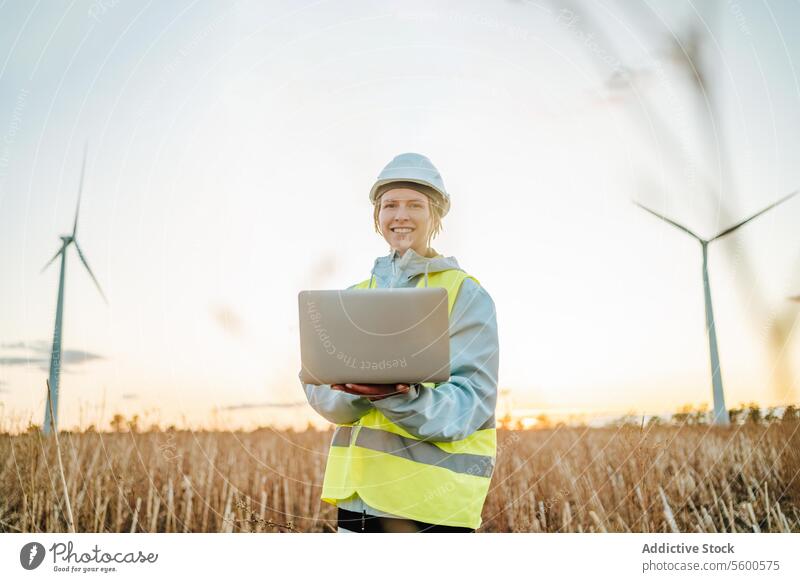 Engineer looking at camera with laptop at wind farm during sunset engineer woman wind turbine field safety gear renewable energy technology work environment