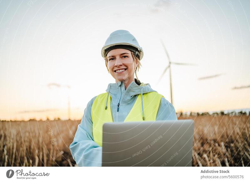 Smiling engineer with laptop near wind turbines at dusk woman field sunset renewable energy safety helmet safety vest technology environment smile professional