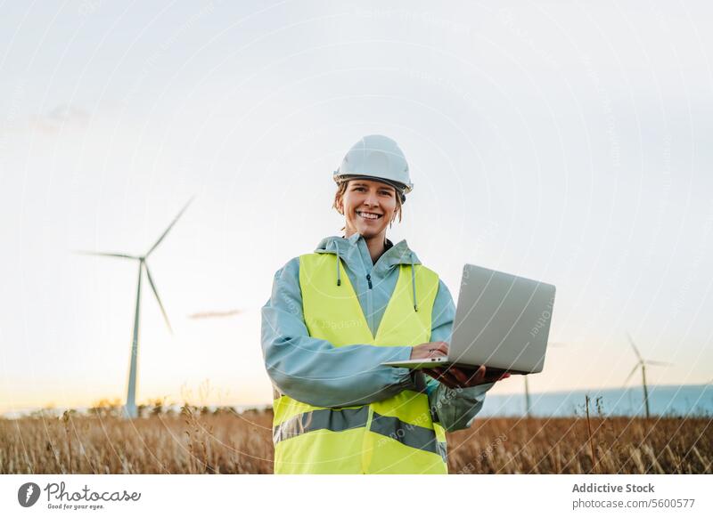 Engineer looking at camera with laptop at wind farm during sunset engineer woman wind turbine field safety gear renewable energy technology work environment