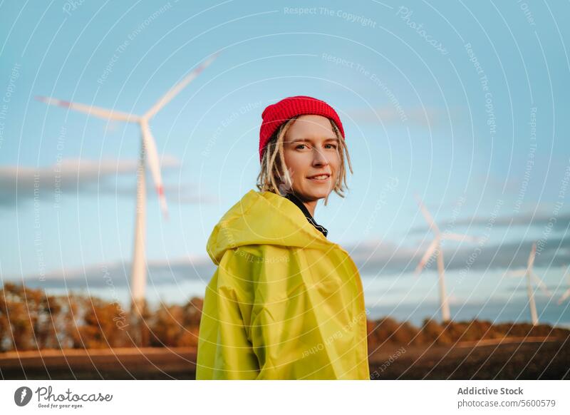 Smiling alternative woman looking at camera in yellow jacket by wind turbines beanie sustainable green energy cheerful young adult portrait dreadlocks renewable