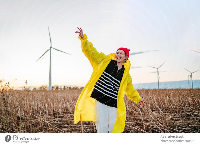 Smiling woman in yellow dancing near wind turbines raincoat red beanie field sunset dance joy vibrant nature renewable energy eco-friendly enjoyment happy fun