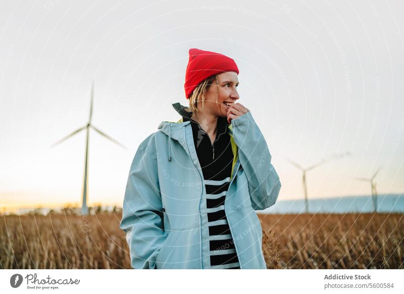 Smiling alternative woman looking away in denim jacket by wind turbines yellow beanie sustainable green energy cheerful young adult portrait dreadlocks