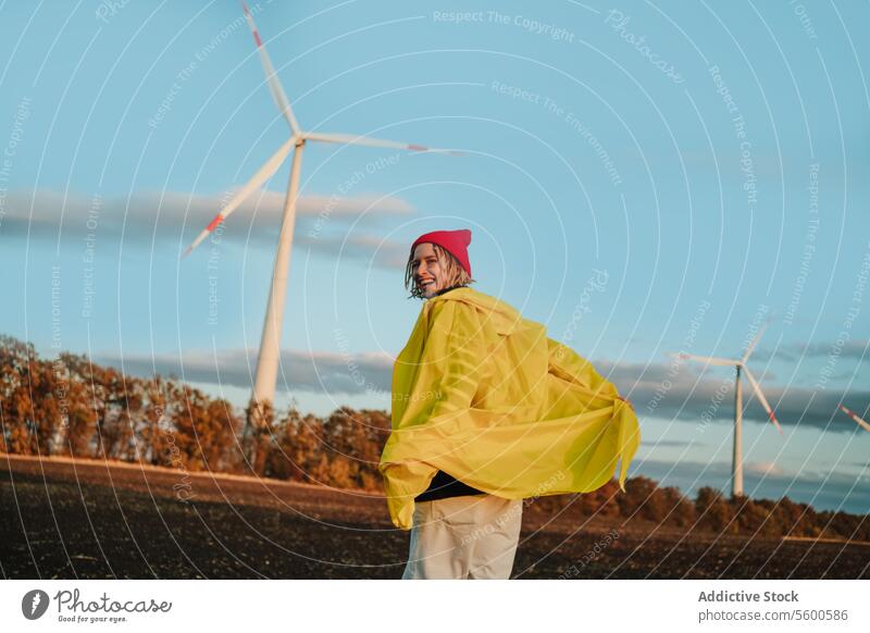 Joyful person in yellow near wind turbines at sunset raincoat pink beanie smile field sky cheerful wind power renewable energy eco-friendly sustainability