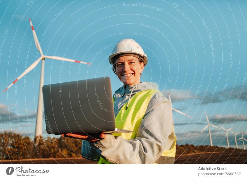 Engineer woman looking at camera with laptop at wind farm during sunset engineer wind turbine safety gear renewable energy technology safety helmet smiling