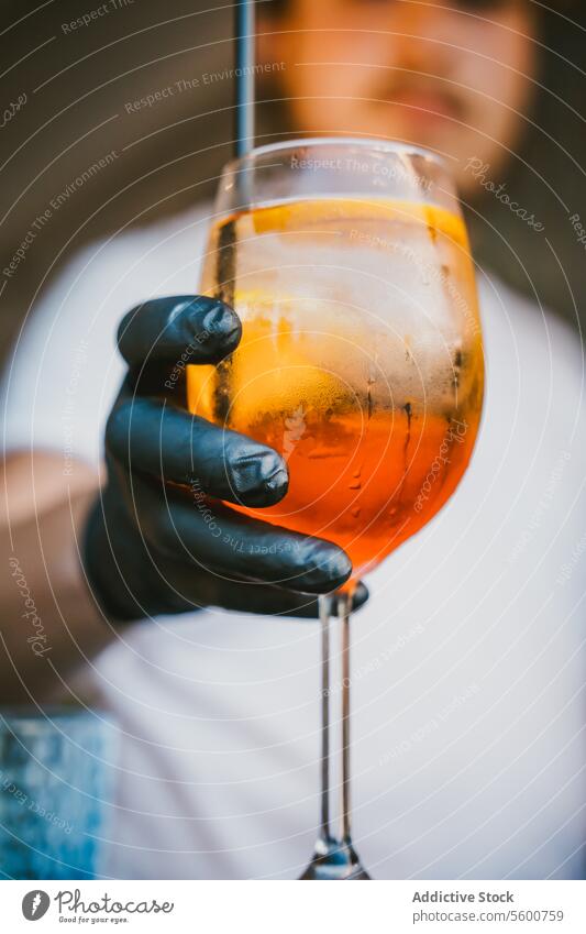 Anonymous bartender with gloves holding an orange cocktail with ice and a straw barman hand beverage alcohol wine glass service mixology close-up drink