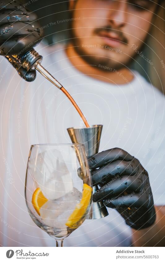 Concentrated young professional barman pouring orange drink in jigger while preparing refreshing cocktail and standing at counter in bar wine glass skill