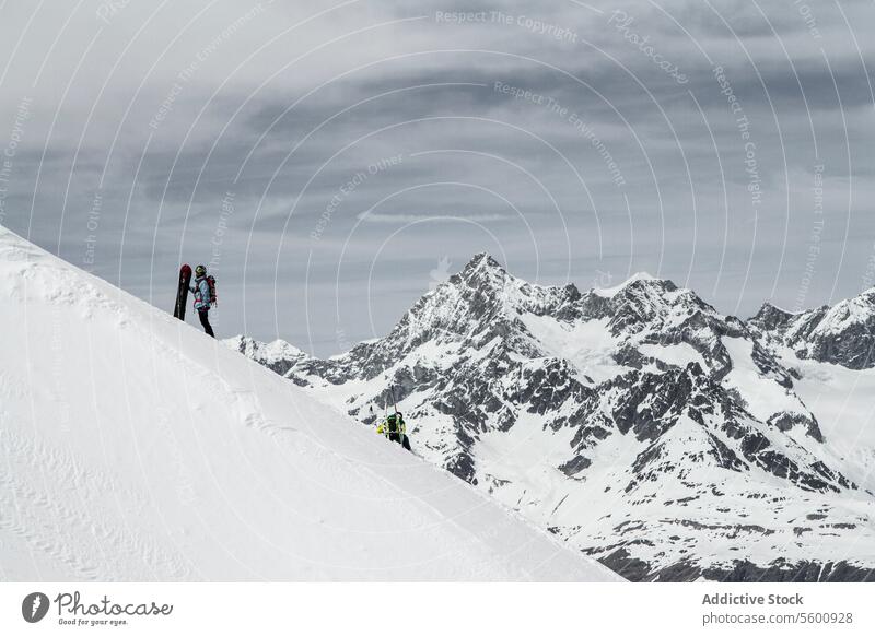 Side view of anonymous climber with snowboard standing on snow covered slope against cloudy sky in winter vacation at Swiss Alps hiker mountain distant