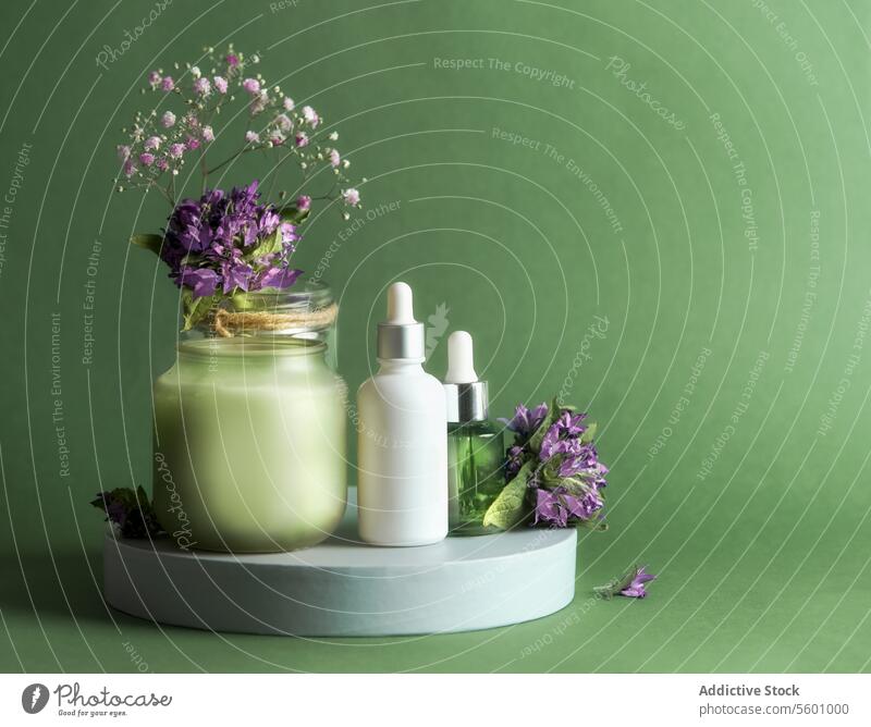 Natural cosmetic product setting with pipette bottle, candle and flowers on podium at green background. Healthy skin care concept with natural products for facial treatment. Front view.