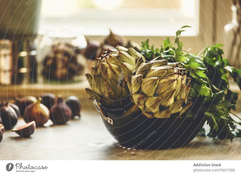 Whole artichokes in bowl with parsley on kitchen table with figs at window background with natural light. Healthy Mediterranean food cooking. Front view. whole
