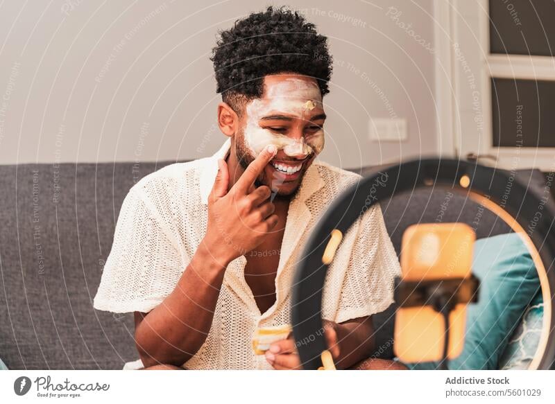 Latin man applies face mask during video blog session skincare home beauty grooming streaming casual mirror ring light couch self-care channel relaxation facial