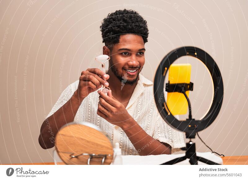 Latin man smiling while recording a video with ring light with a facial roller healthcare skin smartphone content social media young cheerful creator influencer