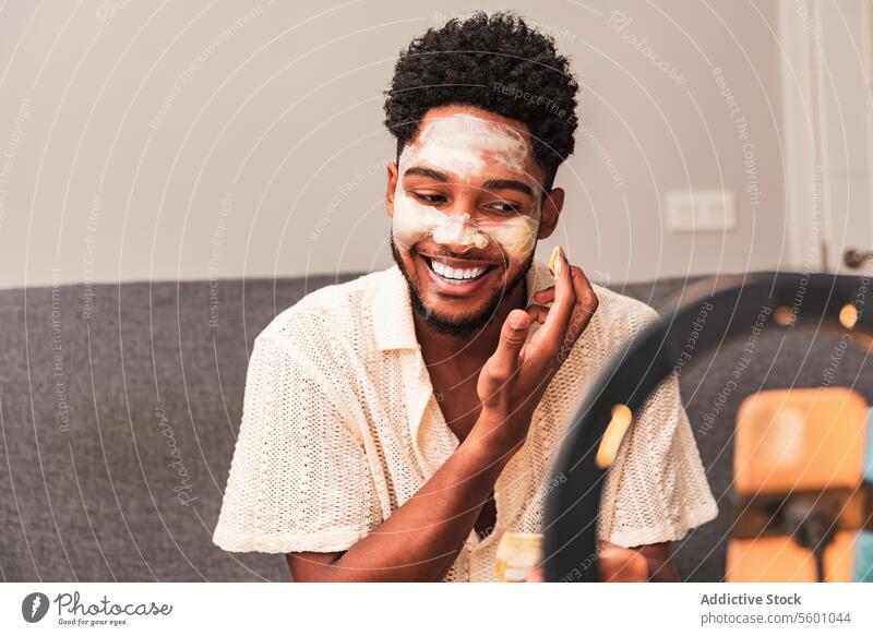 Latin man applies face mask during video blog session skincare home beauty grooming streaming casual mirror ring light couch self-care channel relaxation facial