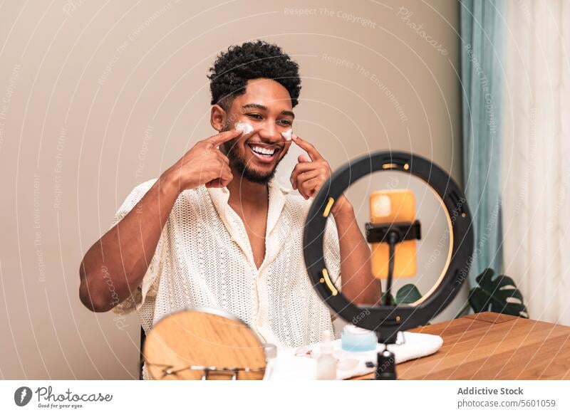 latin man smiles applying skincare routine in mirror home self-care beauty ring light pointing cheek joy happiness grooming personal care facial treatment